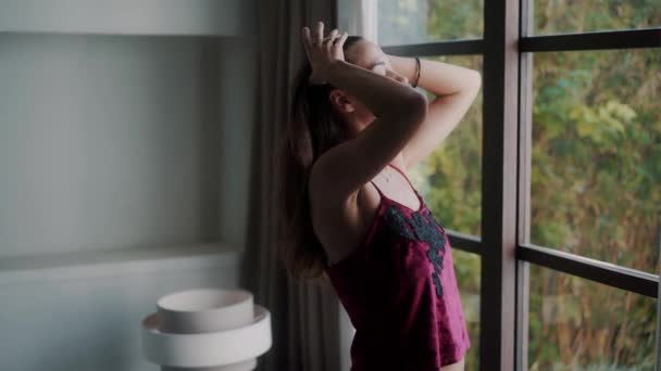 Жінка в піжамі прокидається, насолоджується видом з вікна і зв'язує хвіст у волоссі — стокове відео