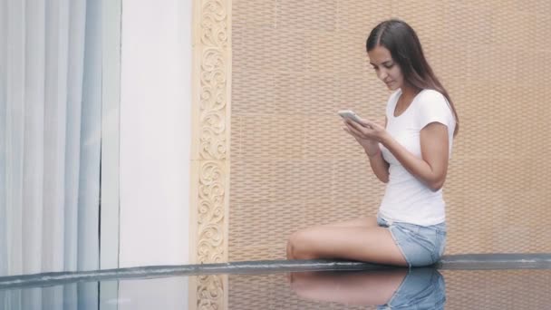 젊은 여자는 수영장 가장자리에 앉아 일하기 위해 전화기를 사용하며, 물에 반사 된다 — 비디오