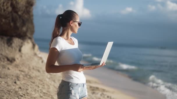 Žena na volné noze ve slunečních brýlích drží notebook, při pohledu na oceán na tropické pláži — Stock video