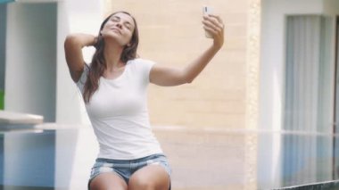 Güzel bir kadının portresi telefonda havuzun kenarında otururken selfie çekiyor.