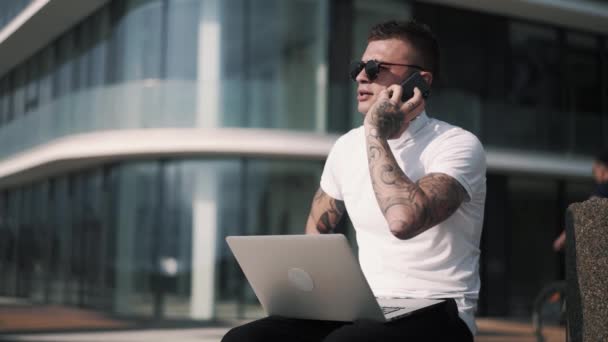 Hombre de negocios tatuado hablando por teléfono y utilizando el ordenador portátil para el trabajo, centro de negocios en segundo plano — Vídeo de stock
