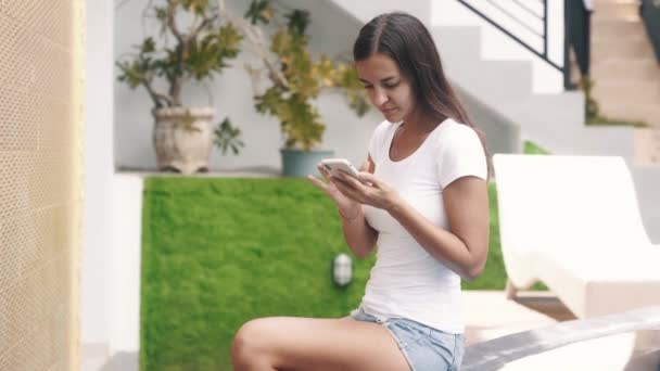 Kobieta freelancer w letnich ubraniach siedzi w pobliżu basenu i używa telefonu na wakacjach — Wideo stockowe