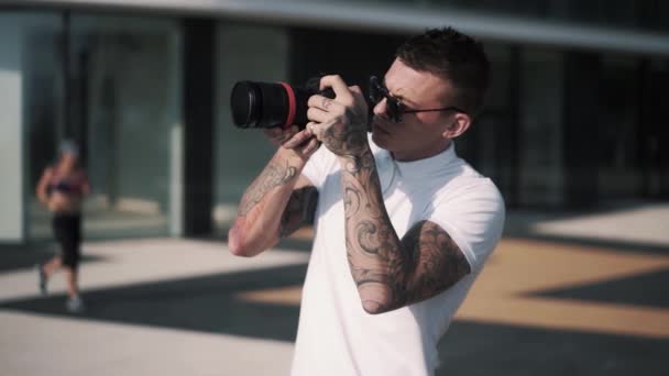 Fotógrafo masculino tatuado en gafas de sol sostiene la cámara en las manos y toma fotos — Vídeo de stock