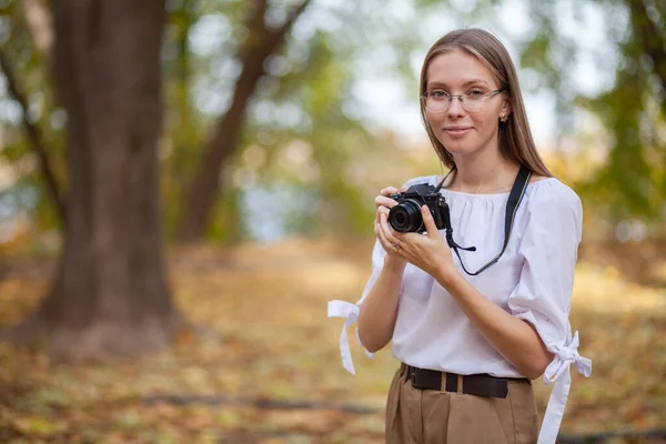 Attractive beautiful young girl holding modern mirror-less camera in autumn park. — Stock Photo, Image