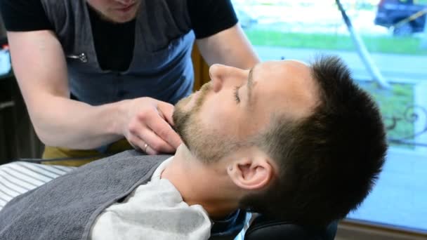 Peluquero corta la barba de los clientes con una recortadora de barba profesional en una barbería sobre fondo claro, primer plano . — Vídeo de stock