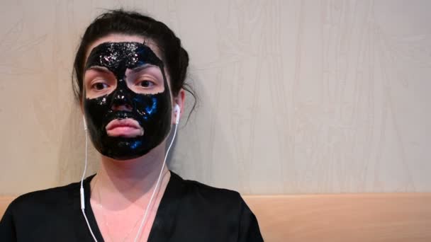 彼女の顔にマスクを浄化し 音楽を聴くと顔面のブラック マスク シートを行う若い女性 — ストック動画