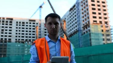 Yetişkin mühendis veya mimar bir tablet işlemi kullanır. Bir ileti yazar veya bir çizim denetler. Arka planı yapıdır. İnşaatçılar beton ve cam modern konut bina inşa ediyoruz.