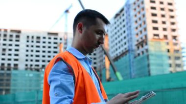 Yetişkin mühendis veya mimar bir tablet işlemi kullanır. Bir ileti yazar veya bir çizim denetler. Arka planı yapıdır. İnşaatçılar beton ve cam modern konut bina inşa ediyoruz.