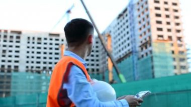 Yetişkin mühendis veya mimar bir tablet işlemi kullanır. Bir ileti yazar veya bir çizim denetler. Arka planı yapıdır. İnşaatçılar beton ve cam modern konut bina inşa ediyoruz.