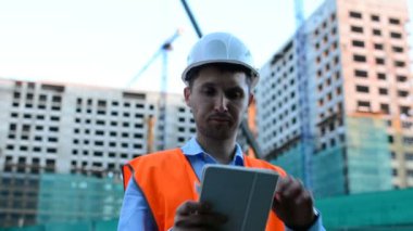 Yetişkin mühendis veya mimar bir tablet işlemi kullanır. Bir ileti yazar veya bir çizim denetler. Arka planı yapıdır. İnşaatçılar beton ve cam modern konut bina inşa ediyoruz.
