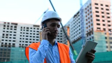 Yetişkin mühendis veya mimar bir tablet işlemi kullanır. Bir ileti yazar veya bir çizim denetler. Arka planı yapıdır. İnşaatçılar beton ve cam modern konut bina inşa ediyoruz.