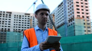 Yetişkin mühendis veya mimar bir tablet işlemi kullanır. Bir ileti yazar veya bir çizim denetler. Arka planı yapıdır. İnşaatçılar beton ve cam modern konut bina inşa ediyoruz.