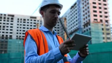 Yetişkin mühendis veya mimar bir tablet işlemi kullanır. Bir ileti yazar veya bir çizim denetler. Arka planı yapıdır. İnşaatçılar beton ve cam modern konut bina inşa ediyoruz.