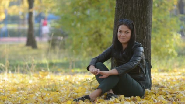 美丽的女孩坐在树下的一棵树在秋天公园微笑 — 图库视频影像