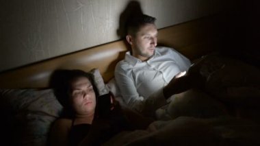 Akıllı telefonları ve tabletleri olan genç bir çift gece geç saatte evde yatıyorlar.