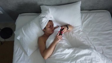 Üst görünümü genç kadın battaniye altında yatakta uyanma. Sevimli kız sabah ilk şey onu akıllı telefon kontrol.