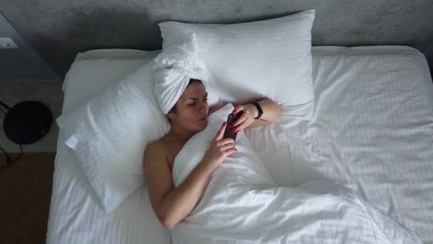 トップビュー若い女性は毛布の下でベッドで目を覚ます かわいい女の子は 朝一番に彼女のスマートフォンをチェック — ストック動画