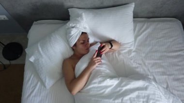 Üst görünümü genç kadın battaniye altında yatakta uyanma. Sevimli kız sabah ilk şey onu akıllı telefon kontrol.