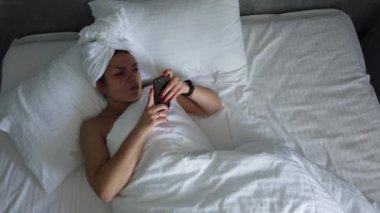 Üst görünümü genç kadın battaniye altında yatakta uyanma. Sevimli kız sabah ilk şey onu akıllı telefon kontrol.
