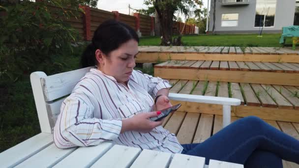 女人使用手机 坐在院子里的长凳上 — 图库视频影像