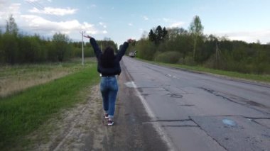Kız boş yolda yürüyor ve otostop çekerken araba yakalamaya çalışıyor.