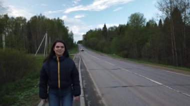 Kız boş yolda yürüyor ve otostop çekerken araba yakalamaya çalışıyor.
