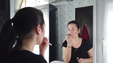 Güzel esmer kadın banyoda aynanın önünde makyaj yapıyor.