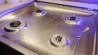 Mavi eldivenli bir adam mutfakta gaz sobasını parlatıyor.