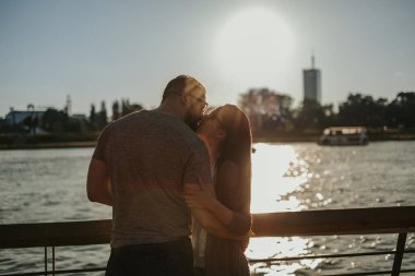 Çift öpüşme ve nehir kıyısında günbatımı, güzel güneşli bir günde sarılma