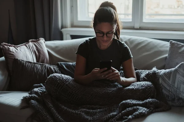 Leende Flicka Med Glasögon Som Sitter Soffan Tittar Mobiltelefon — Stockfoto