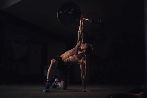 Tattooed, bearded athlete holding weight with one arm in the gym