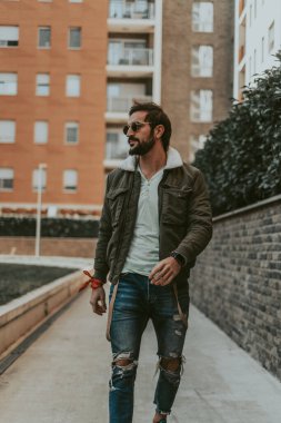 Kendine güvenen, şık, sakallı bir adam şehirde yürüyor.