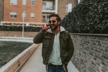 Gülümseyen, sakallı adam eli cebinde telefonla konuşuyor.