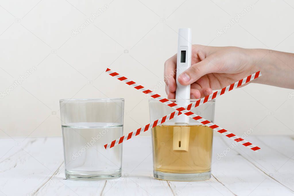 Two glasses with clean and dirty water are on the table. Hand holds the device for the test of water. Red stripes above a glass of dirty water