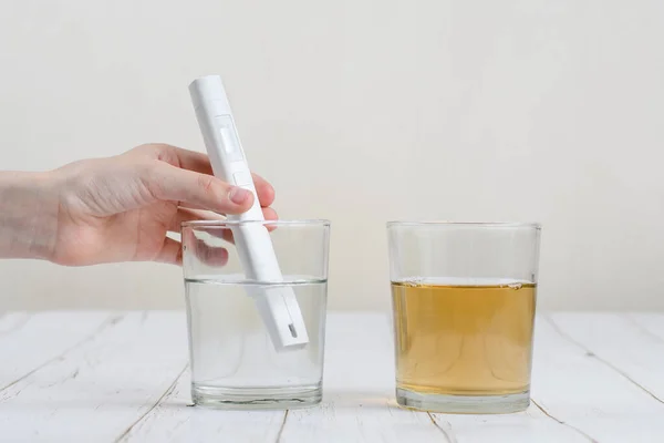 A hand with a device for testing water makes a measurement in a glass with transparent water. Nearby is a glass of dirty water.