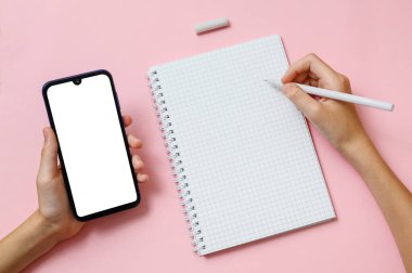 Sağ el bir kalemi bir defter kağıdının üzerinde tutuyor. Sol elinde boş ekranlı bir cep telefonu var.
