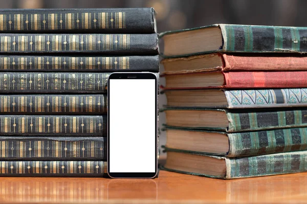 Birçok eski ciltli kitap istiflenmiş. Kitapların yanında boş beyaz ekranlı bir akıllı telefon var..