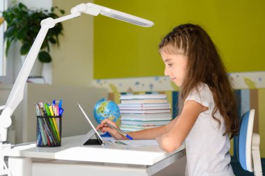Bir kız masaya oturur ve bir tablet kullanır. Kız internet üzerinden uzak dersler alıyor..