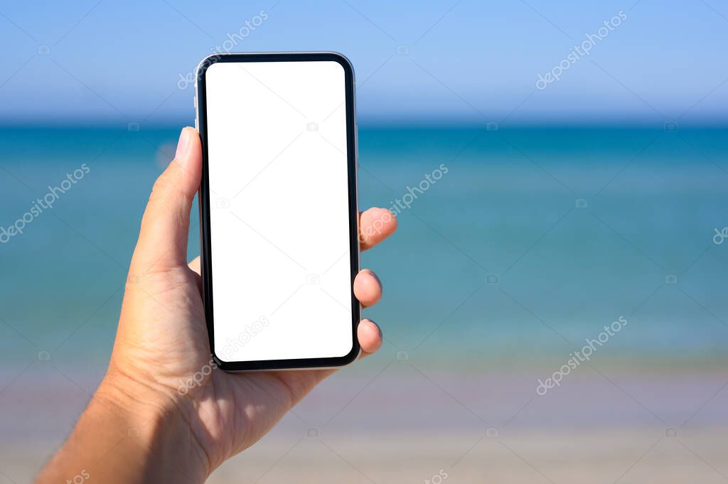 A male hand holds a smartphone with a white display. In the background the sea. Free space for text