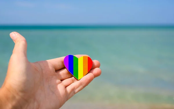 Heart painted in rainbow colors and LGBT flag on the background of the sea. A man\'s hand holds a heart. Vacation concept for LGBT people.