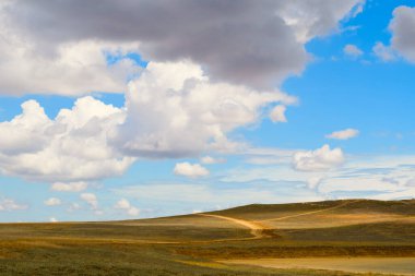 Manzara. Tepeler ve tarlalar üzerinde bulutlarla kaplı gökyüzü. Sahada toprak bir yol var..