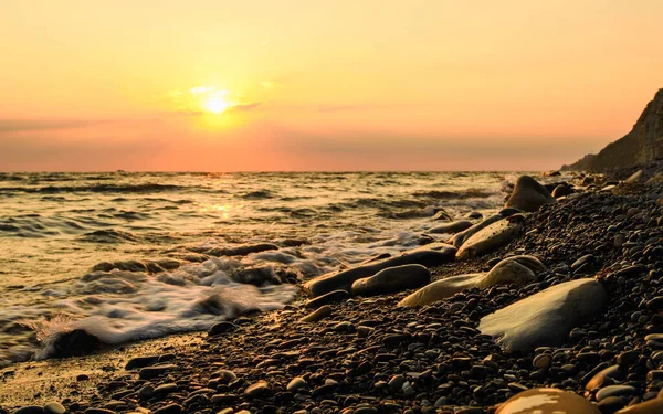 Çakıl taşlı sahilde büyük taşlar. Deniz kenarında gün batımı. Sahilde dalgalar. Uzun süre ateş edildi. Ön plandaki kayalara odaklan..
