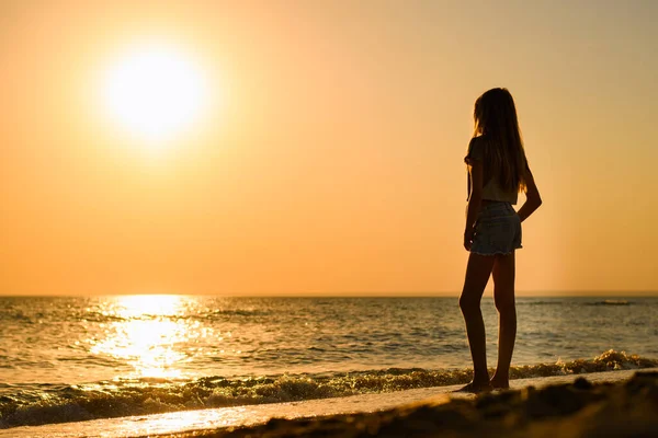 少女は海の近くに裸足で立って夕日を見ている 太陽は海の上に沈む 海の波は砂浜を洗う — ストック写真