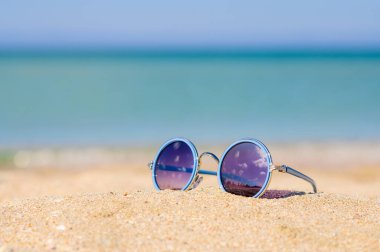 Güneş gözlükleri deniz kıyısındaki kumda yatar. Bulutların yansıması gözlüklerde. Deniz kenarında tatil kavramı.