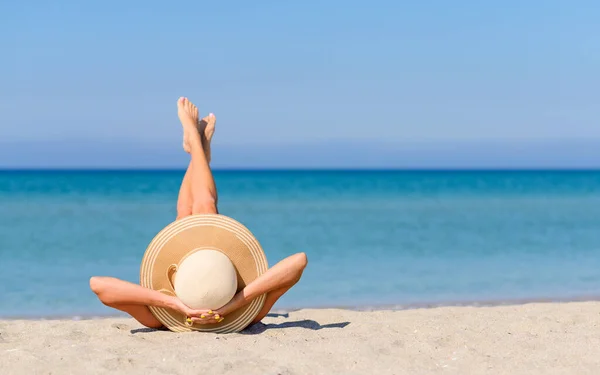 Hasır şapkalı, bacakları ters dönmüş bir kız kumsalda güneşleniyor. Deniz kenarında tatil kavramı.