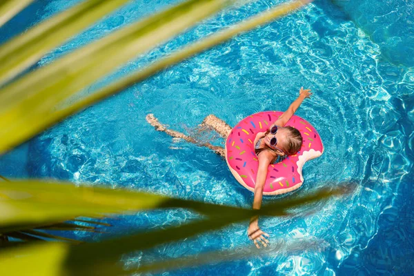 The girl swims in the pool on the circle. In the foreground green leaves of a palm tree