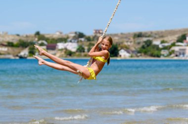 Kız salıncakla denizin üzerinde uçuyor. Deniz kenarında tatil yapan neşeli bir kızın portresi..