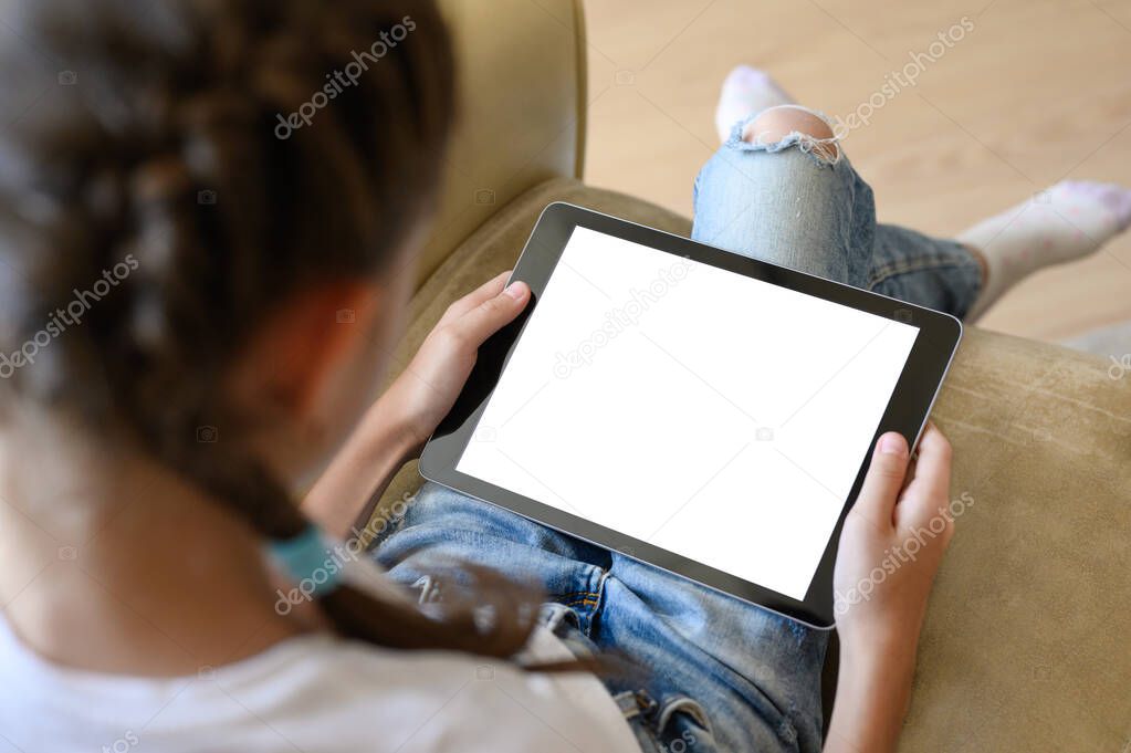 A tablet with a white display in the hands of a girl. The girl sits at home on an armchair and plays on the tablet.