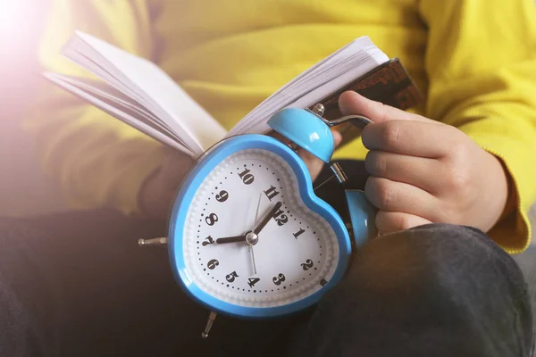 Un niño con una chaqueta amarilla sosteniendo un libro abierto y un despertador en sus manos —  Fotos de Stock