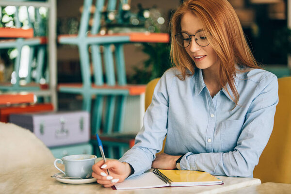 Red-haired girl in a cafe. Is drinking coffee. Checks reports at the computer.